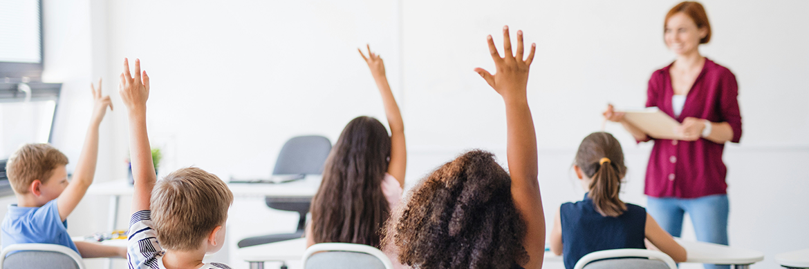 Group of students raising their hands during a classroom discussion with a female teacher standing in front of the class
