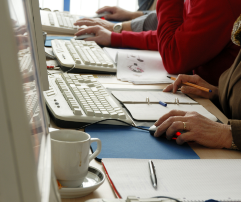 There people in a computer class sitting at desktop computers. You can see the arms of the people, which are on the right. The keyboards are on the left. 