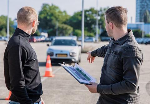 photo of two men turned to the side, one is point at a car with a clipboard