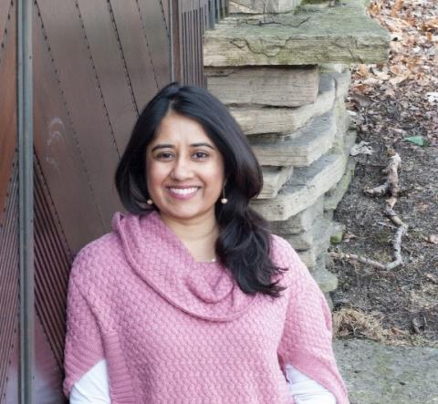 photo of a smiling woman in a pink sweater with dark black hair leaning against a wooden door