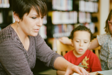 Woman pointing to a book with two children sitting beside her in the library