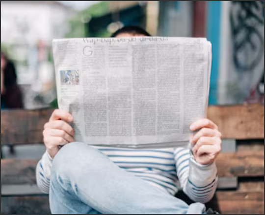 Library News linked image showing a person holding a newspaper in front of their face while sitting on a bench