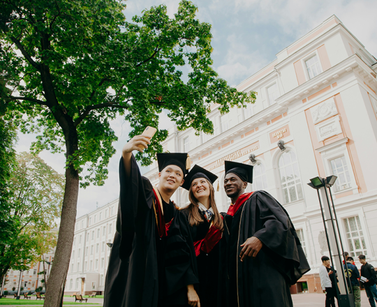 Three teens, two boys and a girl, smile and pose for a selfie in graduation gowns and ca[s
