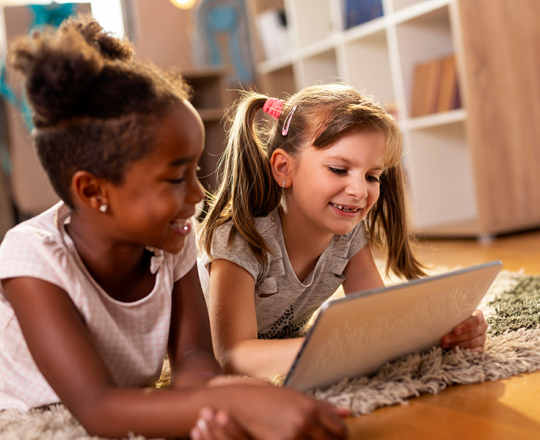 Two girls smiling while laying on the floor with a tablet in front of them